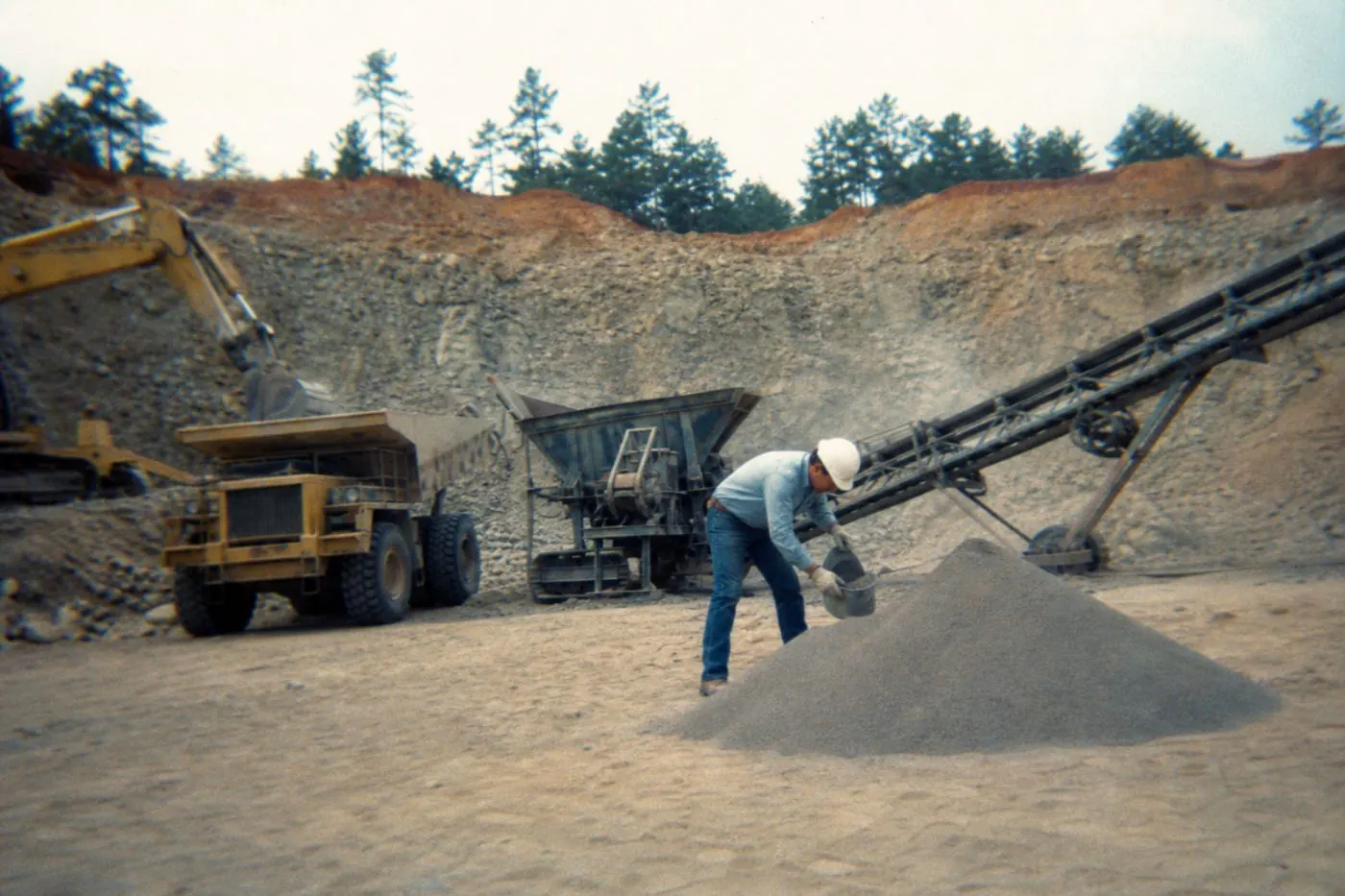 A worker in a hard hat scooping a sample from a granule stockpile at a quarry