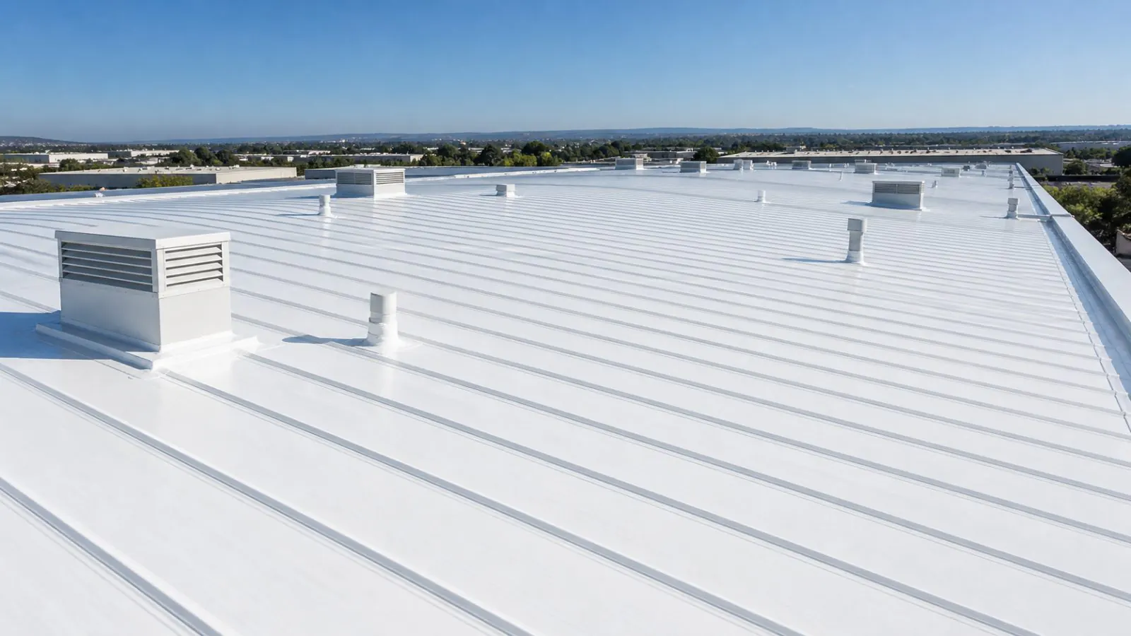 A white-coated standing-seam metal roof with vents and curbs under a clear sky