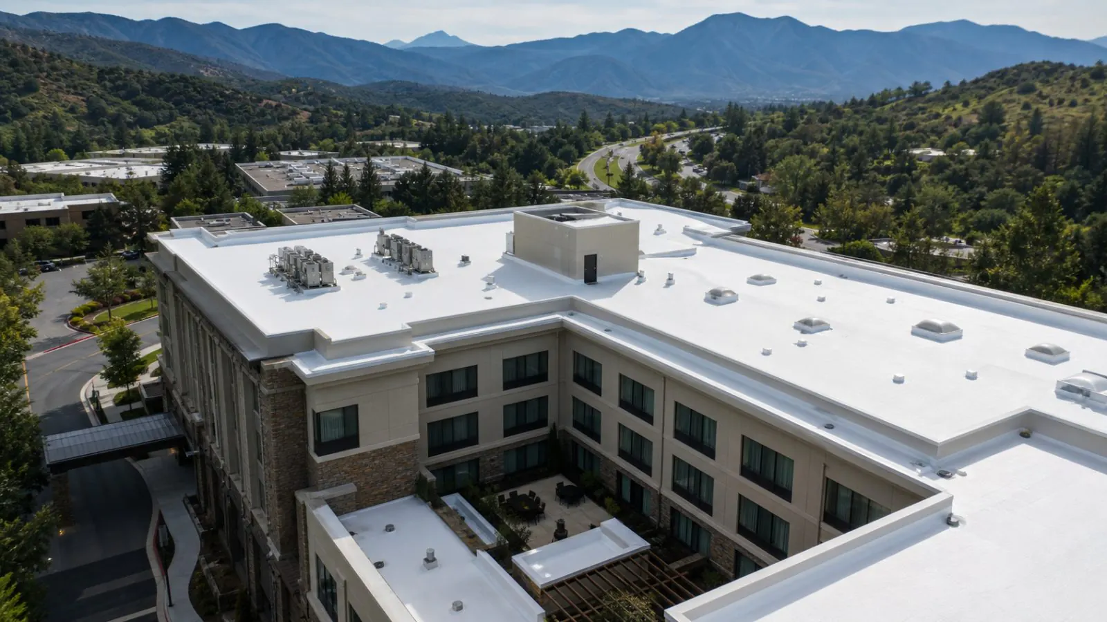 A white-coated flat roof on a multi-level building with wooded hills behind it, seen from above